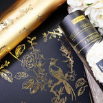 Load image into Gallery viewer, Decorative paper with gold floral and bird designs on a black background, next to a roll of gold paper and a container labeled 're design'.
