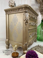Load image into Gallery viewer, Dotted Flourish Redesign with Prima Stencil Delicate Filigree Damask Crest Pattern raised stencil on the side of a gold metallic sideboard
